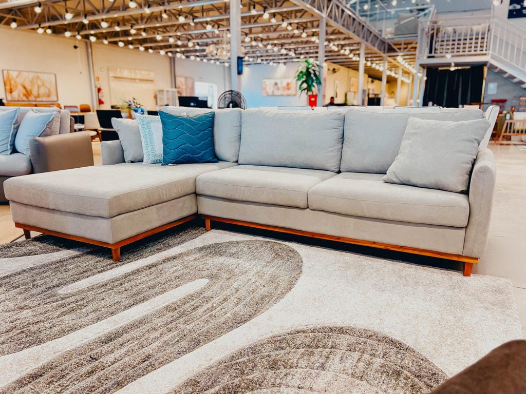Sofá de Living Lyon com Chaise Fixa – Vista Frontal Completa Visão frontal do Sofá Lyon cinza claro em uma loja, com chaise no lado esquerdo, pés de madeira aparente e almofadas decorativas em tons de azul e cinza sobre um tapete ondulado.