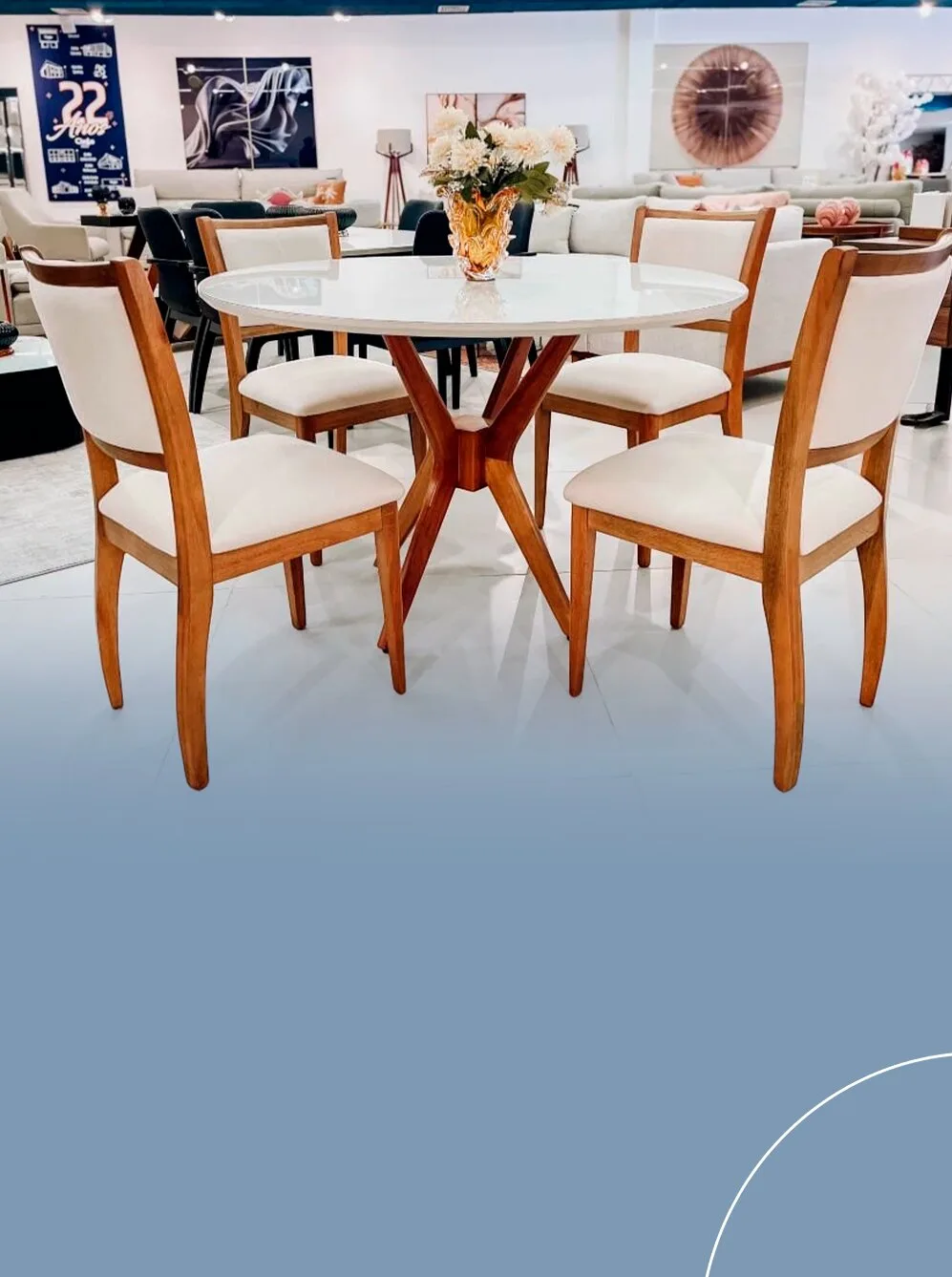 Conjunto de mesa de jantar redonda com tampo branco e base central de madeira cruzada, acompanhada de quatro cadeiras de encosto alto estofadas em tom off-white.